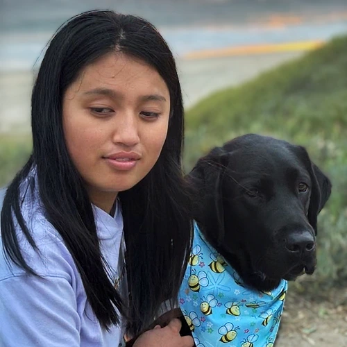 Lynn Wu, a woman with a medium complexion and long dark hair, sitting outdoors beside her guide dog Busby, a black Labrador wearing a blue bumble bee-patterned bandana.