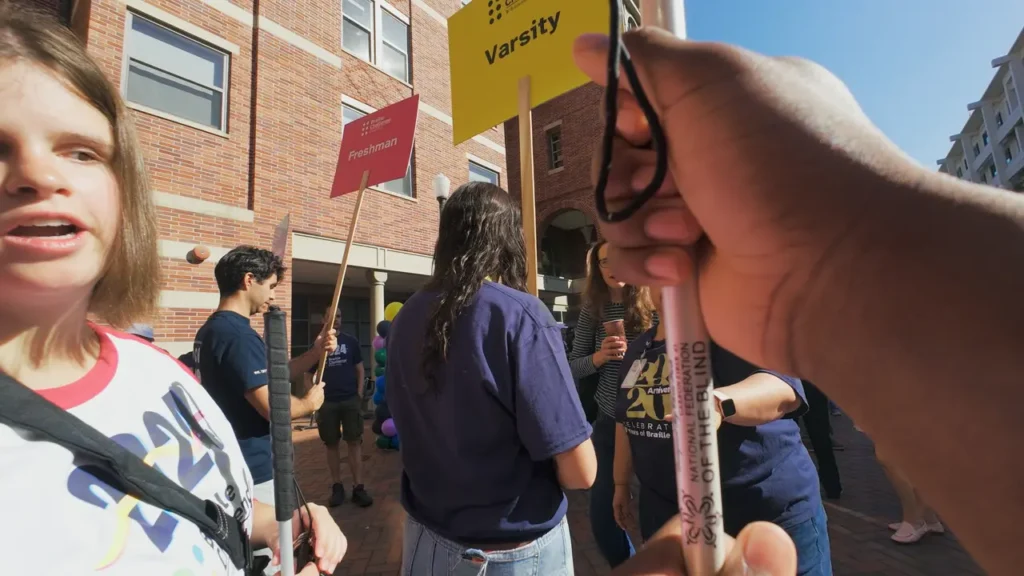A group of people gather outdoors in a brick courtyard, holding colorful signs labeled with team names like “Varsity” and “Freshman.” A hand holding a white cane is visible in the foreground, and a young woman with a cane stands on the left, mid-conversation.