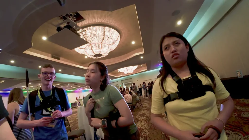 Three young people wearing chest-mounted cameras stand in a ballroom under large chandeliers, talking and waiting with others milling around in the background.
