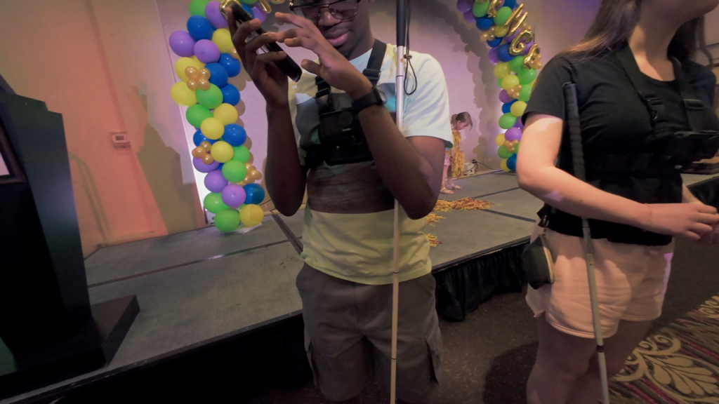 Two teens with white canes and chest-mounted cameras stand near a stage decorated with colorful balloon arches, while a young child plays with confetti in the background.