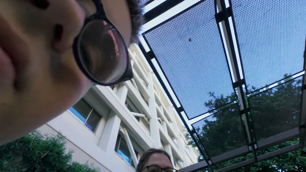 A close-up of a boy wearing glasses' face partially entering the frame, with another person visible below and a metal canopy and building behind them.