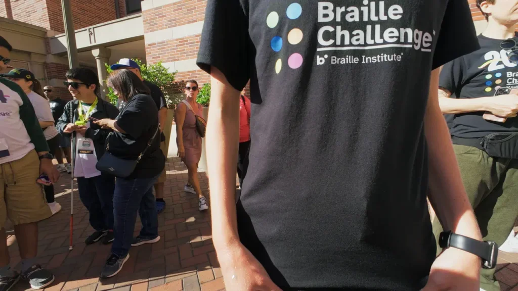 A person wearing a black “Braille Challenge” T-shirt stands in the foreground while groups of participants and families gather and talk in the sunny brick courtyard behind them.