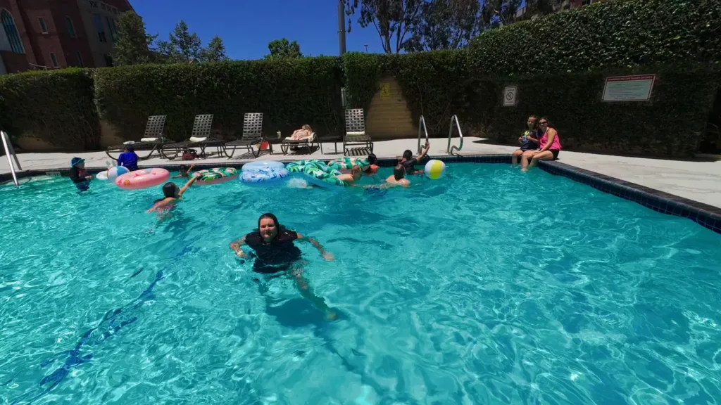 Kids and teens swim in a bright outdoor pool with floaties and a beach ball, while a few people sit along the pool’s edge in the sun.